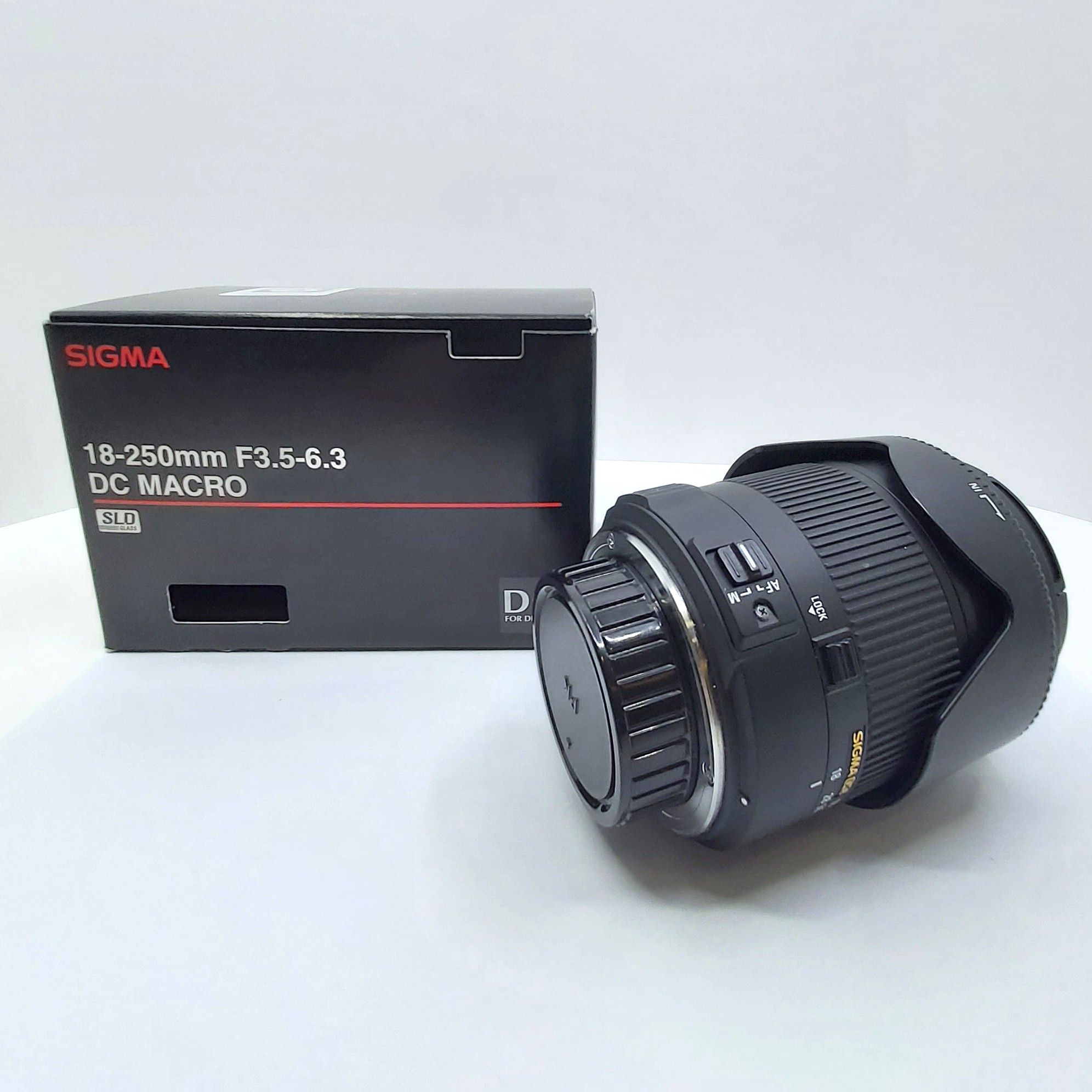 Sigma 18-250mm F3.5-6.3 DC Macro Objektiv für Pentax liegt mit Softtasche vor schwarzer Originalverpackung, Nahaufnahme zeigt Details wie Fokusschalter und Bajonett, ideal für Allround- und Makrofotografie.