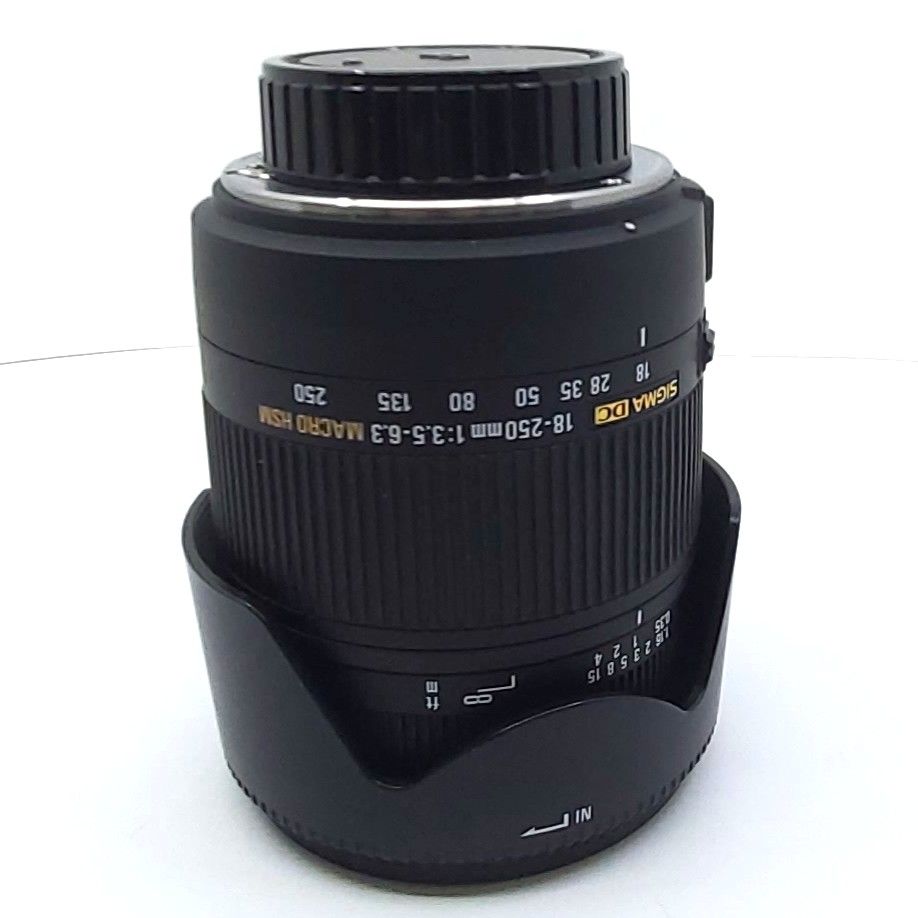Sigma 18-250mm F3.5-6.3 DC Macro Objektiv für Pentax, schwarzes Zoomobjektiv mit gelber Beschriftung, aufrecht stehend mit abnehmbarer Gegenlichtblende, inklusive Softtasche und Originalverpackung, Produktfoto auf weißem Hintergrund.